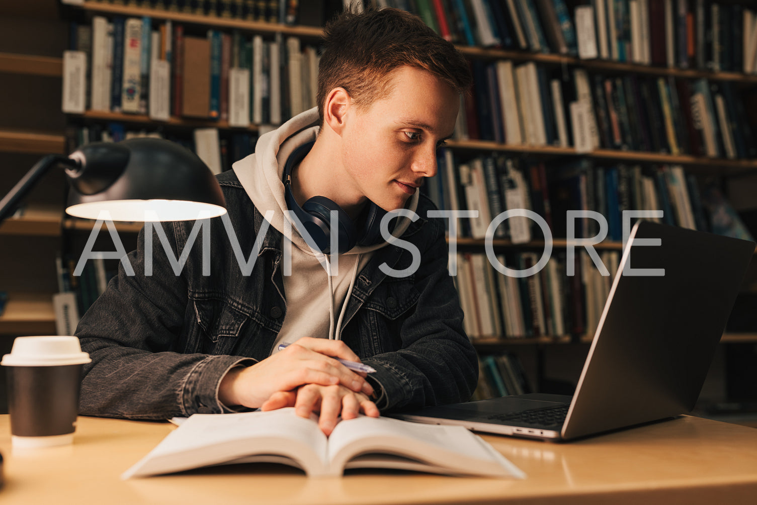 Male student in library looking at laptop. Young man sitting at desk preparing exams at evening.