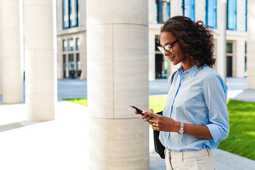 Portrait of modern businesswoman standing outdoors and texting on cell phone