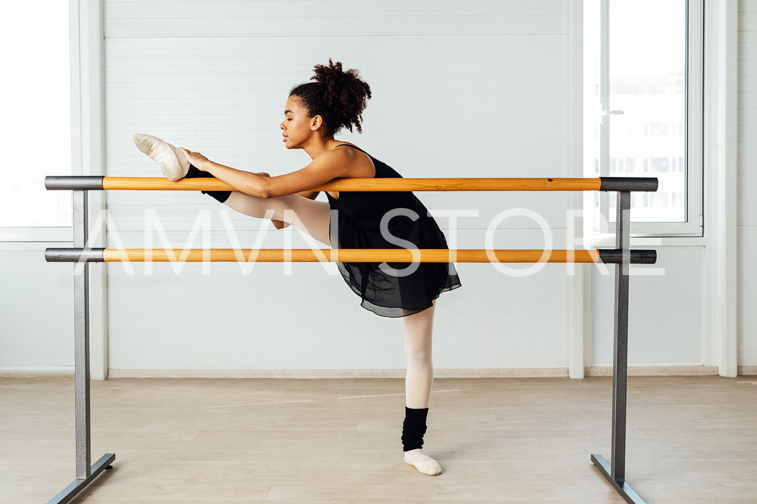 Female ballet dancer with black hair stretching her leg. Side view of young woman preparing for a ballet class in studio.	