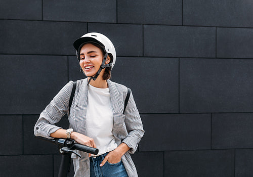 Stylish woman laughing with closed eyes while standing outdoors with electric scooter