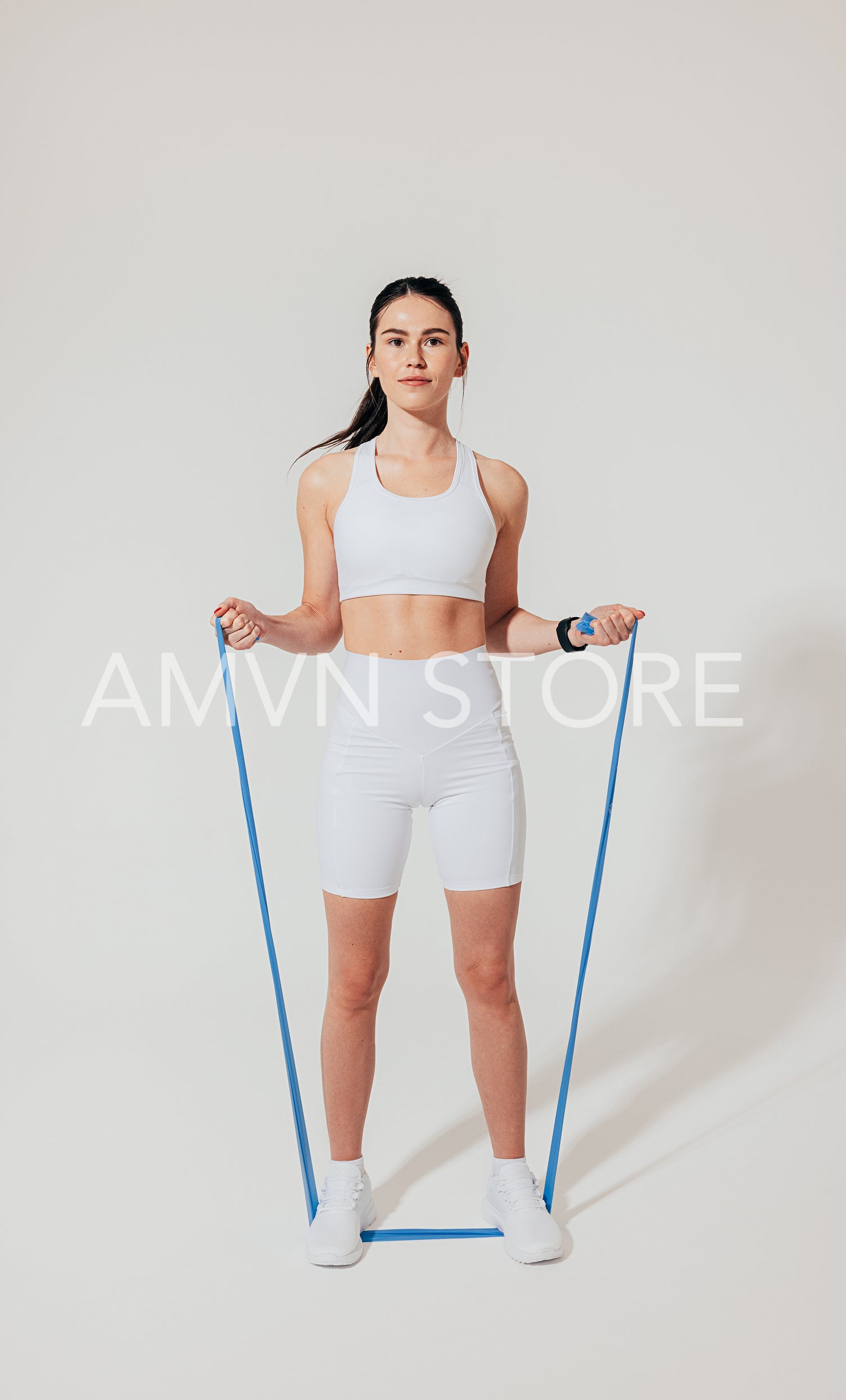 Full length of a young female in white fitness clothes practicing with a resistance band on a white backdrop