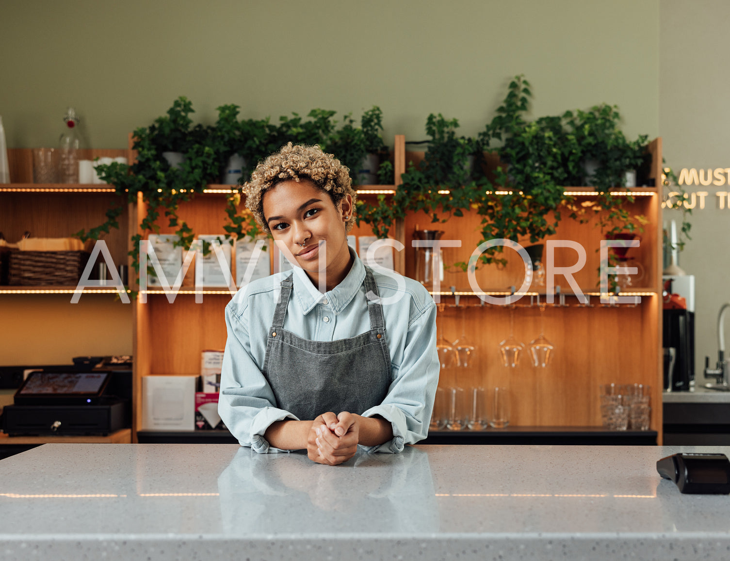 Barista in an apron with short curly hair leaning on a counter. Coffeeshop owner looking at camera.