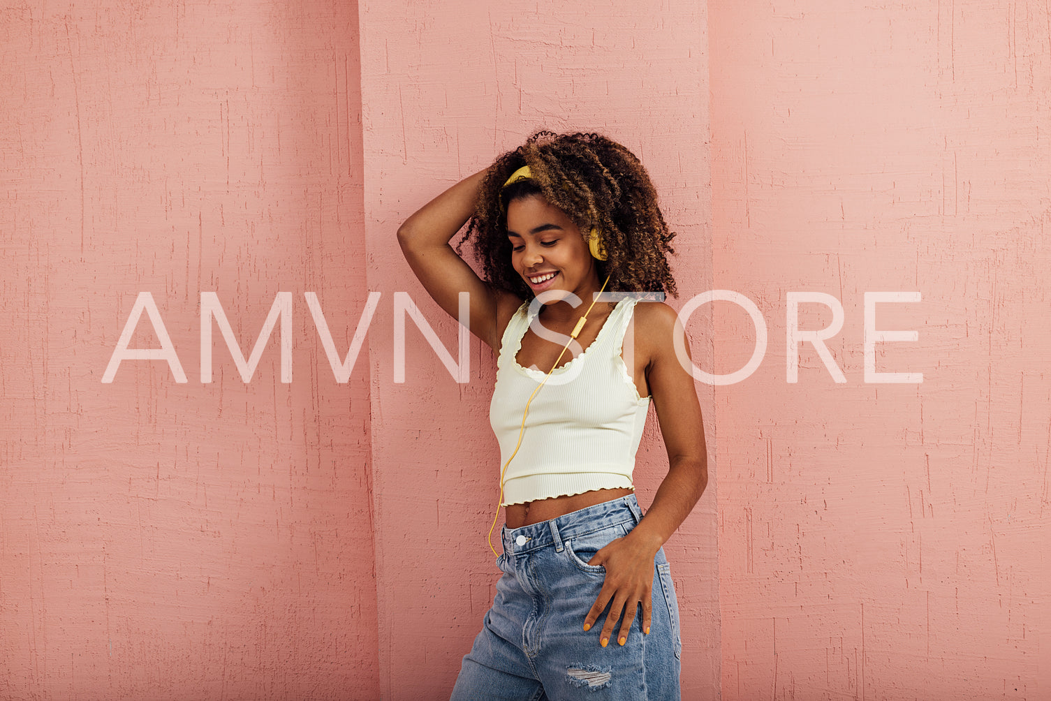 Cheerful woman with curly hair leaning pink wall while listening music