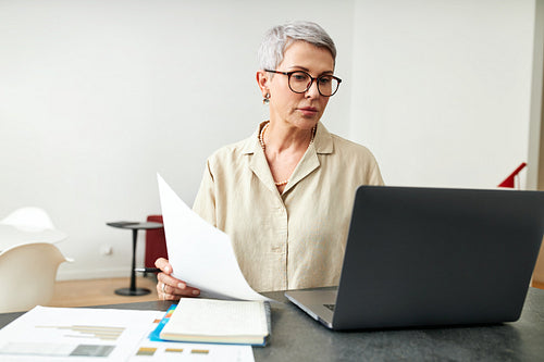 Mature woman with documents using laptop at desk in living room