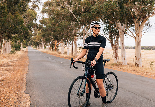 Female cyclist equipped in professional outfit relaxing outdoors. Sportswoman standing on empty road with pro bike.