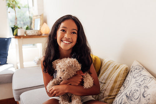 Portrait of a happy girl sitting on a sofa in living room hugging teddy bear