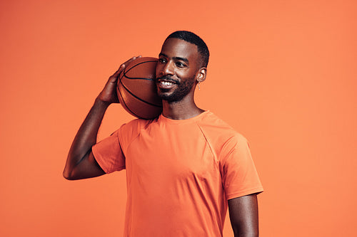 Smiling athlete with basket ball on his shoulder standing in a studio and looking away