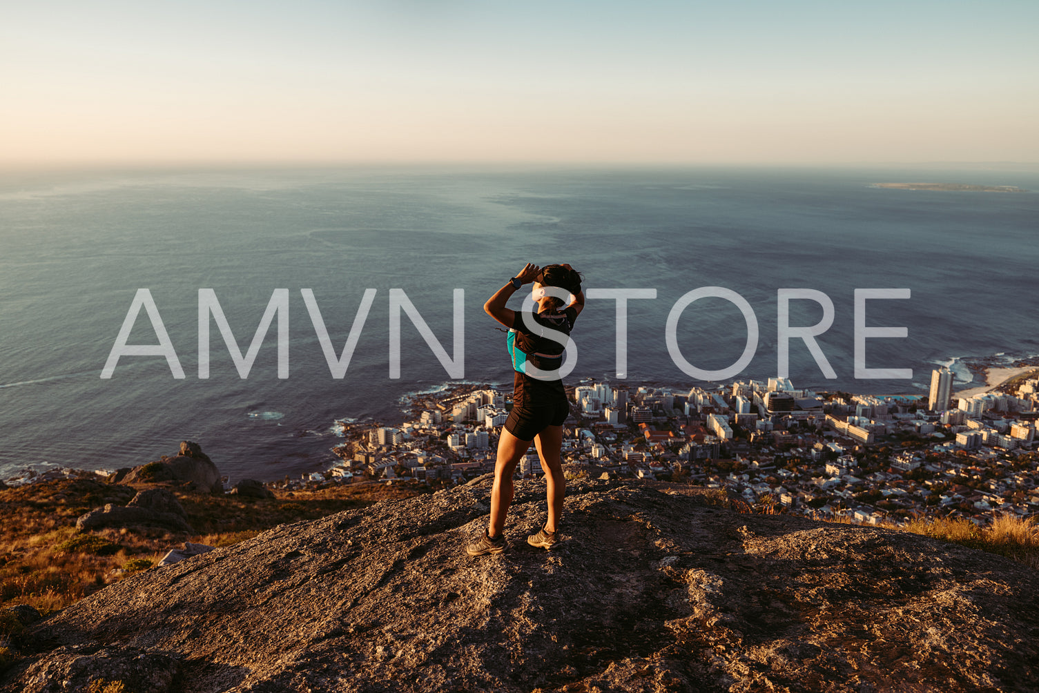 Woman in sportswear standing on the cliff and looking at the view at sunset. Female trail runner relaxing on mountain top looking at the ocean.