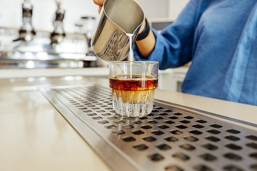 Unrecognizable barista pouring milk from a stainless steel milk jug into a glass for flat white