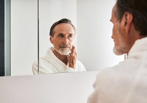 Mature man in a white bathrobe looking at a mirror in bathroom at morning