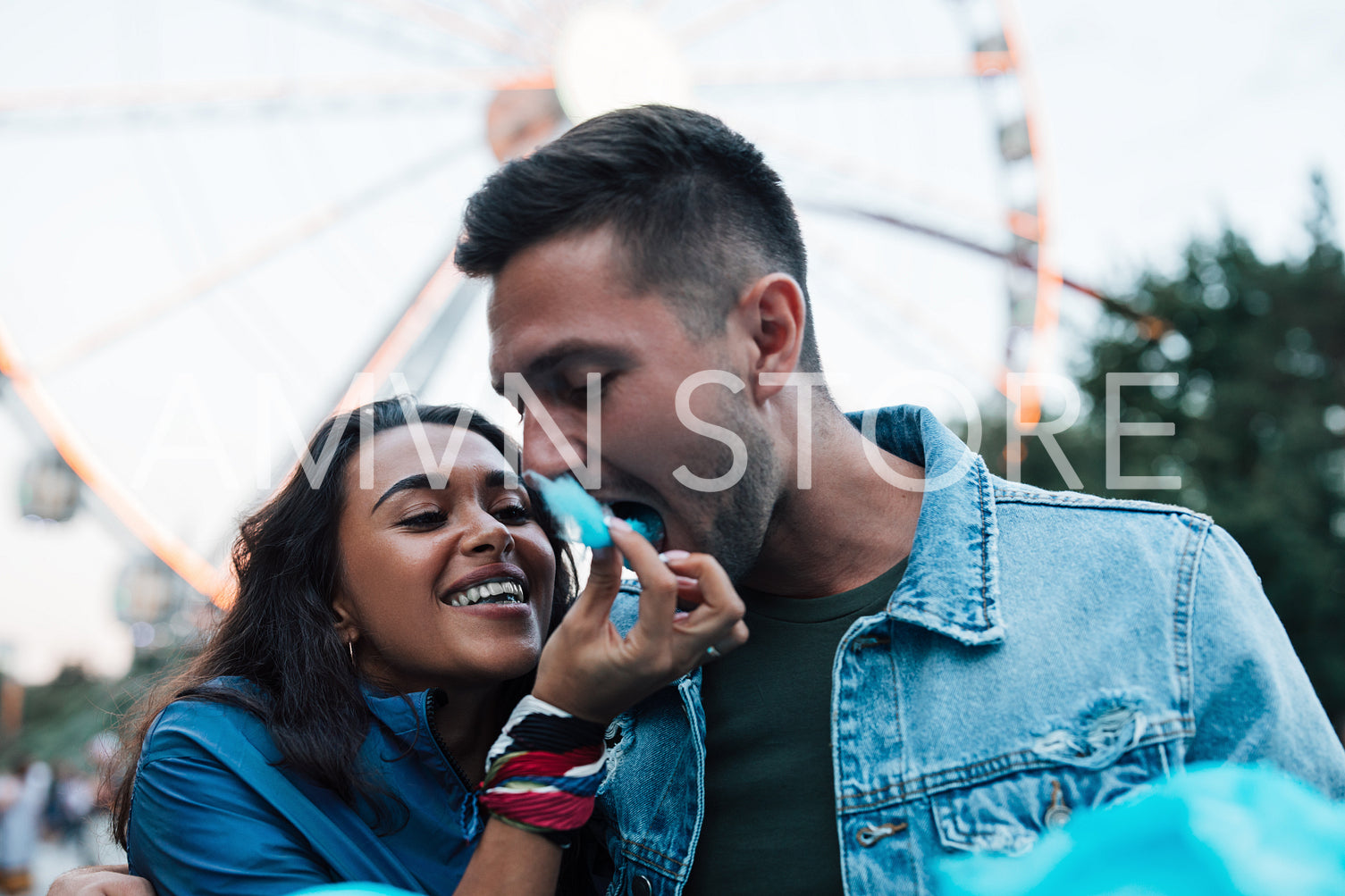 Young couple eats cotton candy during the festival. Cheerful girlfriend feeds her boyfriend with blue cotton candy.