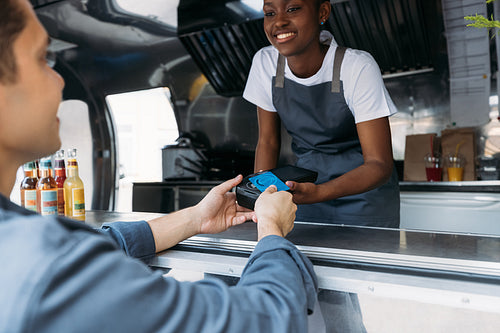 Customer paying with a credit card at a food truck. Smiling saleswoman in apron holding a pos terminal while buyer making NFC transaction.