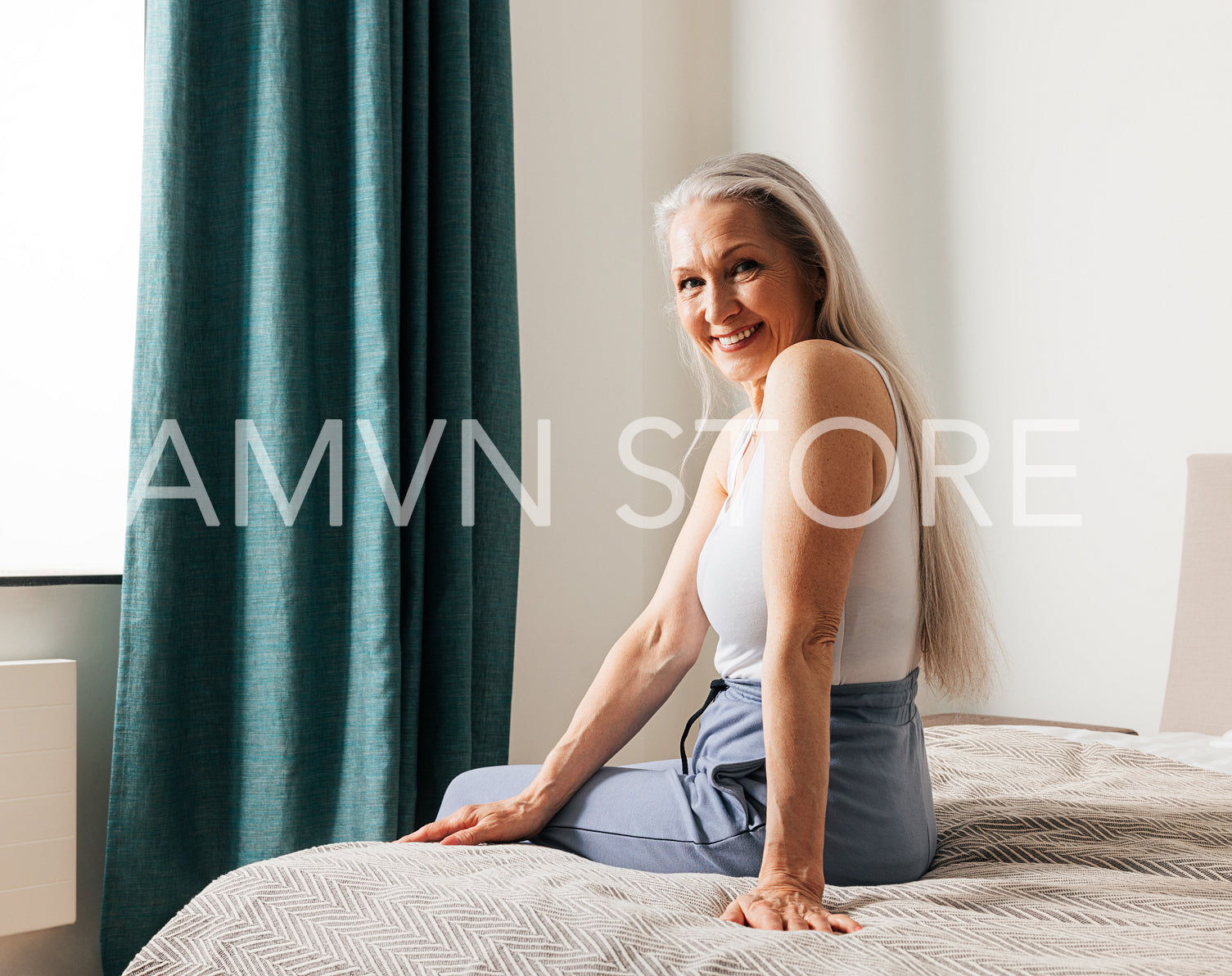 Smiling senior woman with long white hair looking at the camera while sitting on a bed. Female in casuals in bedroom.