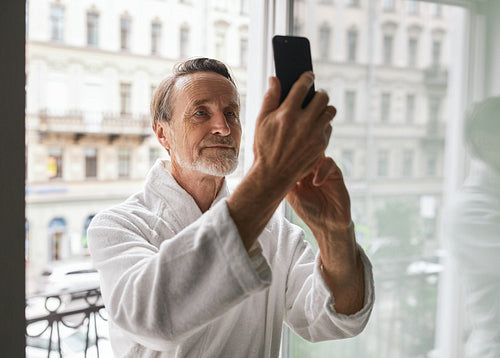 Senior man wearing bathrobe standing on balcony of a hotel room and taking a selfie