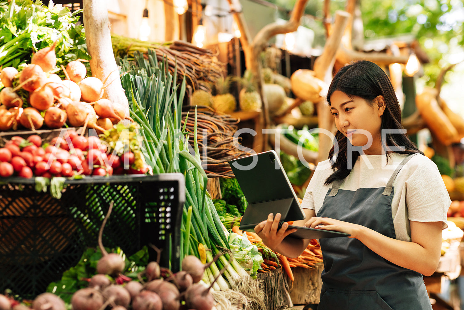 Young businesswoman in an apron working at a local food market. Female with digital tablet at a stall with vegetables.