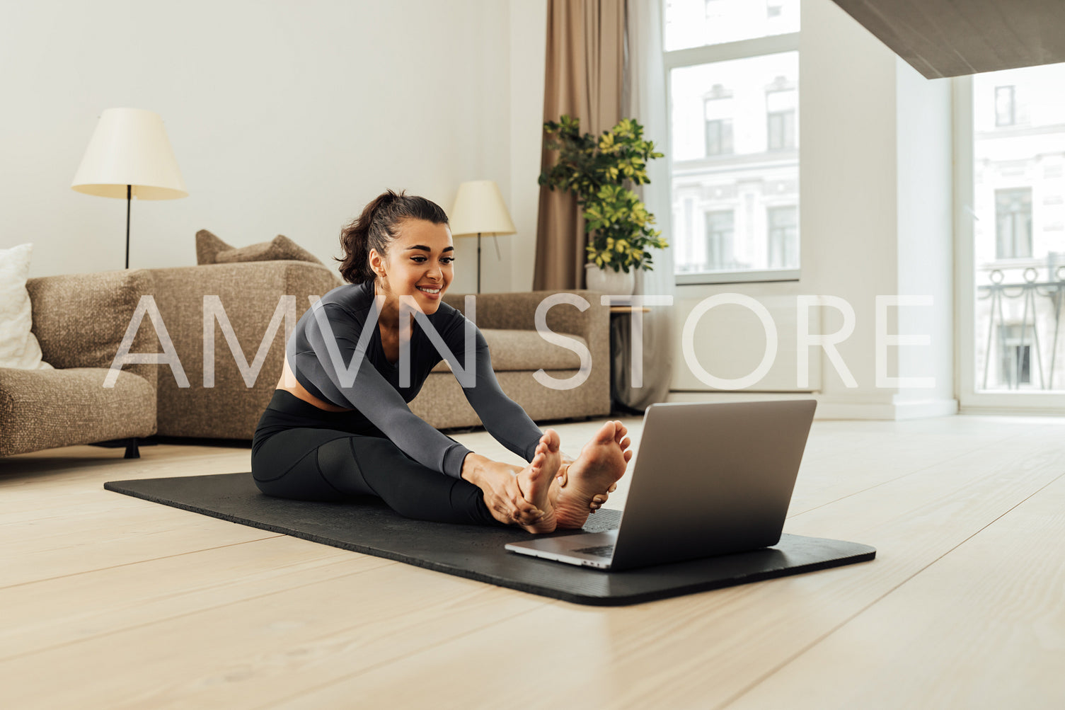 Smiling woman in sportswear sitting on a mat in front of a laptop and stretching her body	