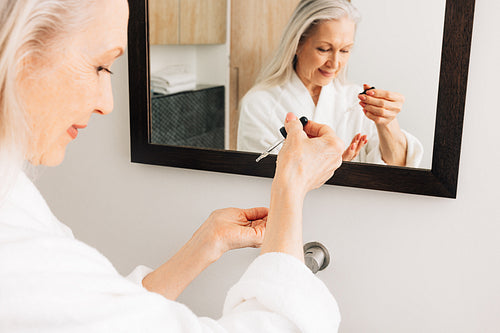 Senior female in a bathrobe drips liquid facial treatment on a palm in front of a mirror. Hands of an aged woman with a hyaluronic dropper.