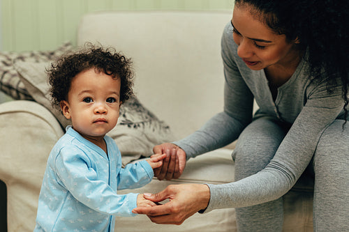 Young mother holding her child hands in living room. Toddler looking at camera.