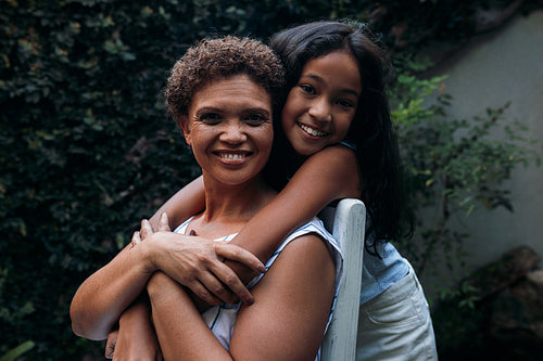 Portrait of a girl and her grandmother. Granny sitting on a chair granddaughter hugging her from back.