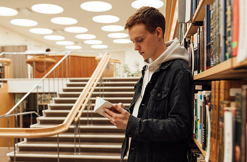 Side view of a male student standing at bookshelf and reading