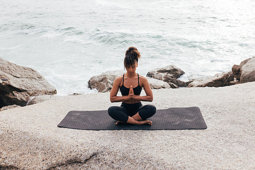 Woman in praying pose sitting on mat with closed eyes by ocean