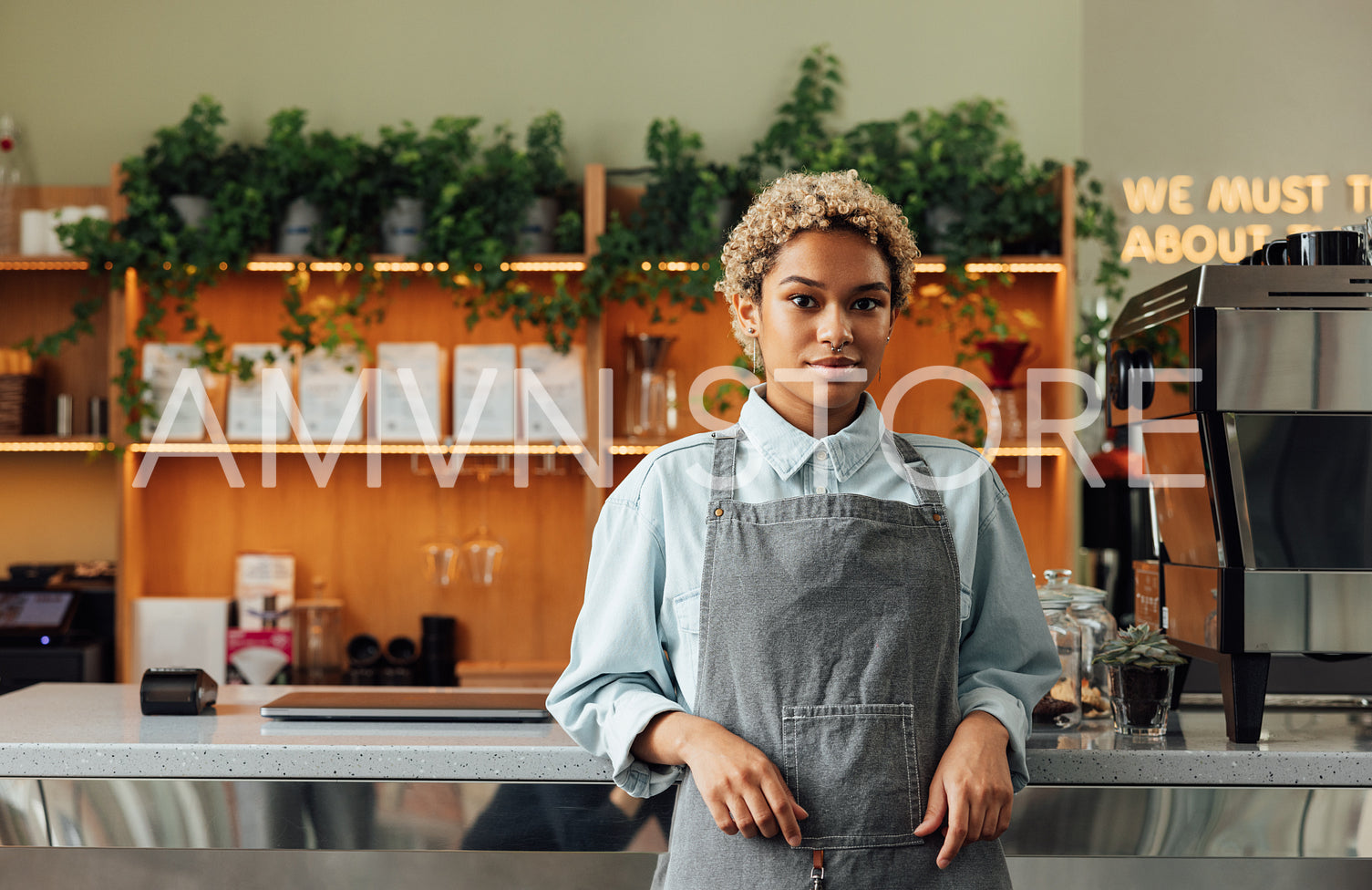 Confident female barista leaning on a counter looking at camera. Young woman in an apron working as a barista standing in a cafe.