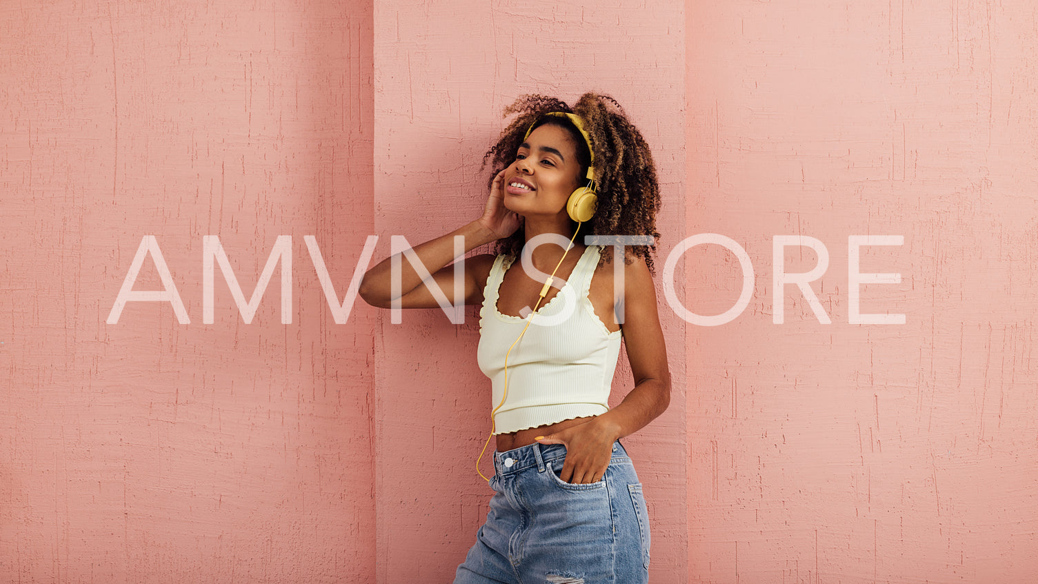 Young female in casuals leaning on a pink wall listening to music by yellow headphones