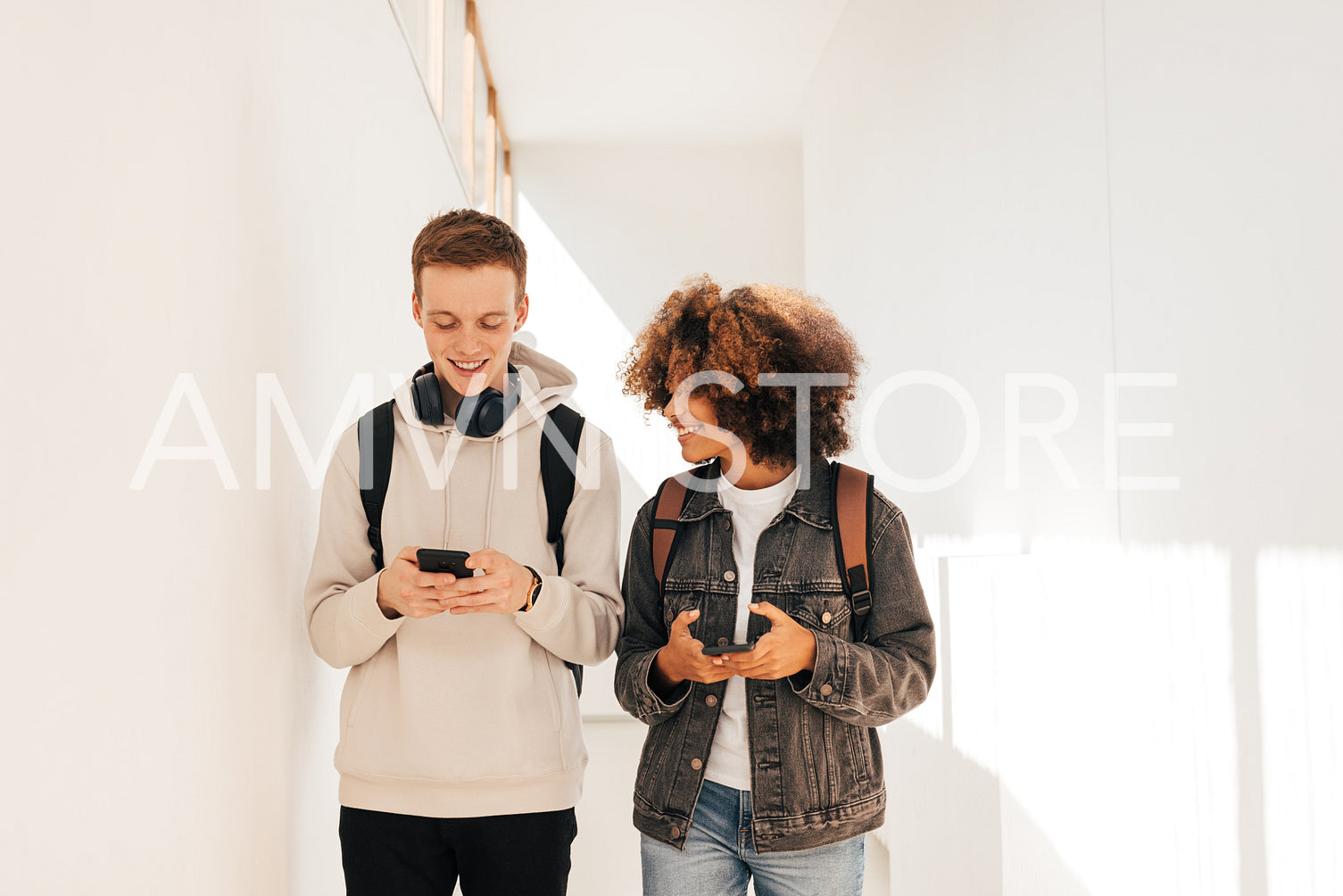 Two smiling students walking in corridor and chatting while holding smartphones