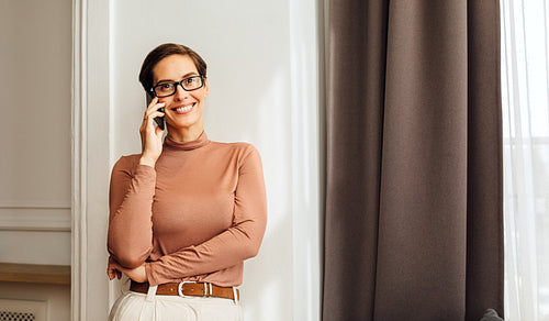 Smiling businesswoman standing at wall in apartment and talking on mobile phone