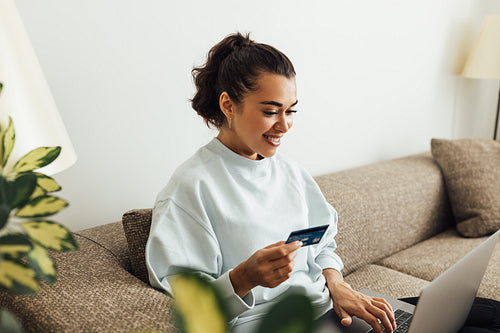 Smiling brunette woman doing online payment. Young female holding her credit card while typing on laptop.