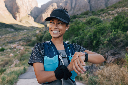 Smiling woman hiker in cap looking away. Female trail runner checking smart watch while standing in valley.