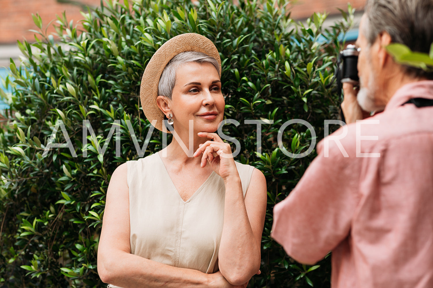 Smiling senior woman in a straw hat looking at the camera while her husband photographs her