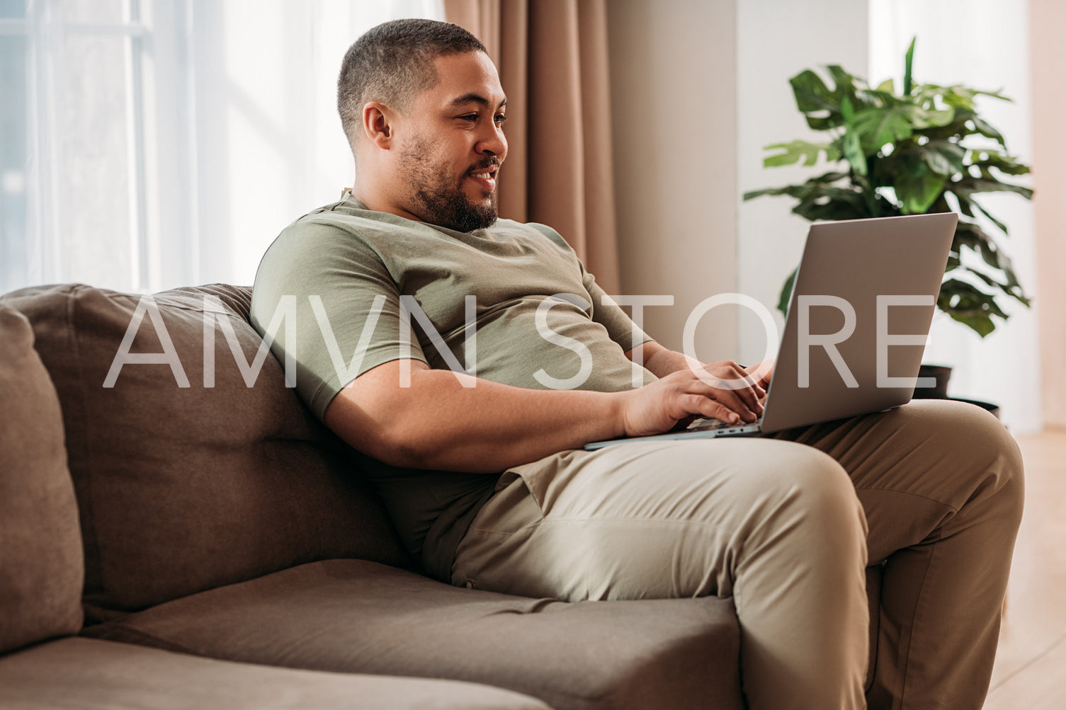 Smiling man with laptop on his hips sitting on a sofa. Young male working from home.	