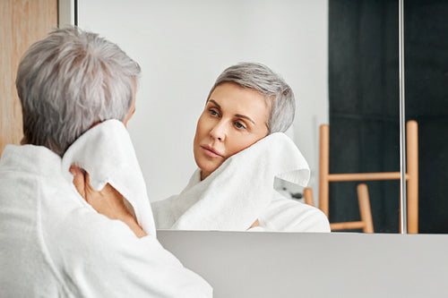 Mature woman with short grey hair looking at her reflection in bathroom mirror while wiping cheek