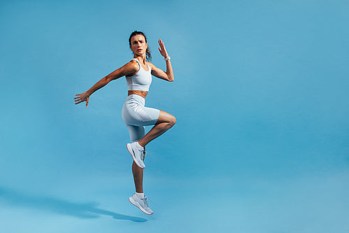 Slim woman jumping in studio on blue background. Muscular female in fitness clothes exercising.