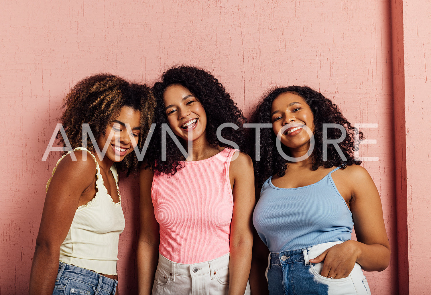 Portrait of a three smiling women with curly hair posing together against pink wall looking at camera