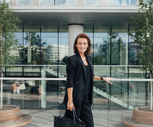 Smiling businesswoman in black formal wear holding a bag standing against an office building. Middle-aged female standing outdoors.