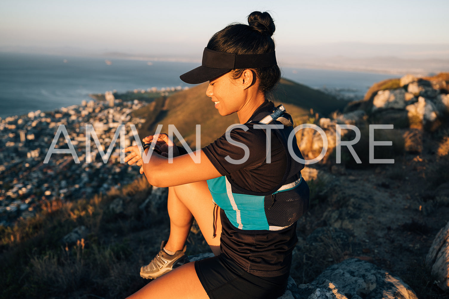 Sportswoman in cap sitting on hill checking fitness tracker. Smiling female looking at smartwatch after hike.