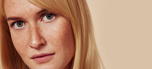 Close-up studio shot of a young beautiful female with naturally smooth skin. Cropped studio shot of woman with freckles looking at camera over beige backdrop.