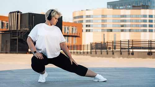 Plus size woman stretching her leg. Young curvy female exercising on roof.