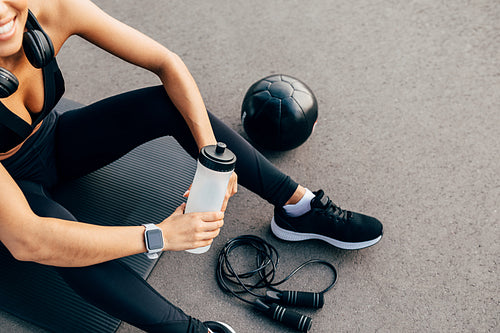 Cropped shot of unrecognizable sport woman sitting on a mat outdoors and holding bottle