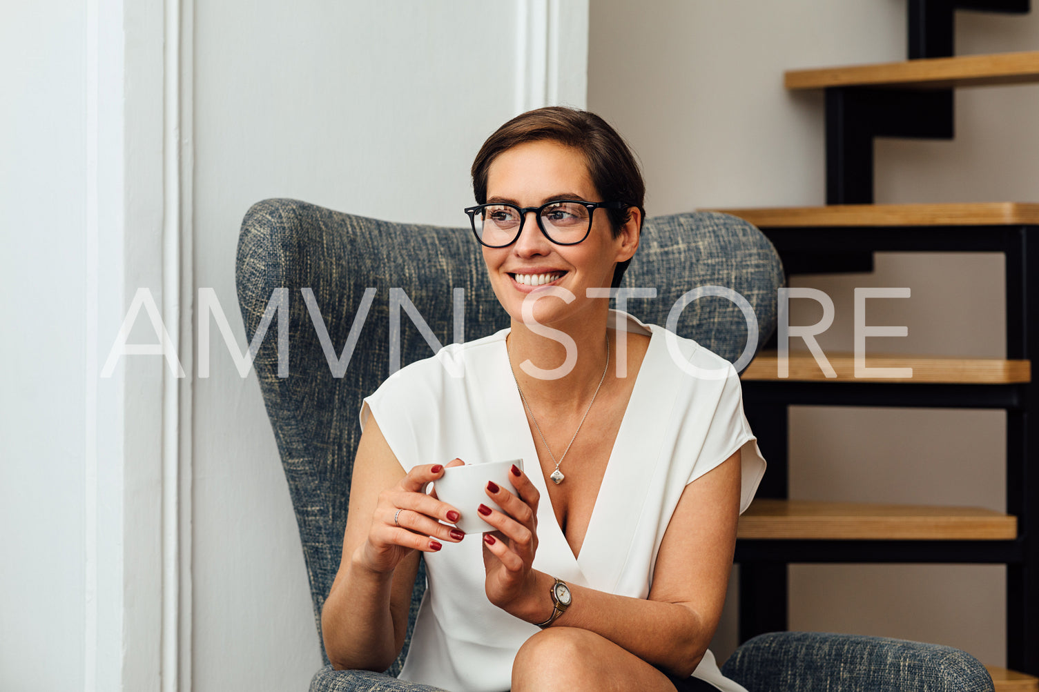 Smiling woman in eyeglasses holding a cup and looking away while sitting in apartment on an armchair	