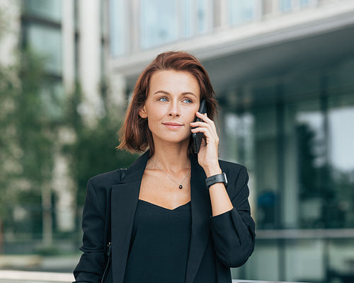 Middle-aged businesswoman in formal clothes making phone call. Portrait of a young adult female with ginger hair talking on a mobile phone while standing outdoors.