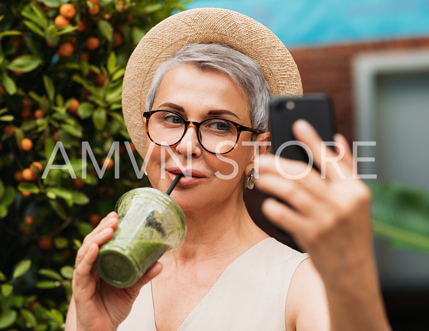 Aged woman in eyeglasses drinks smoothie and taking selfie outdoors