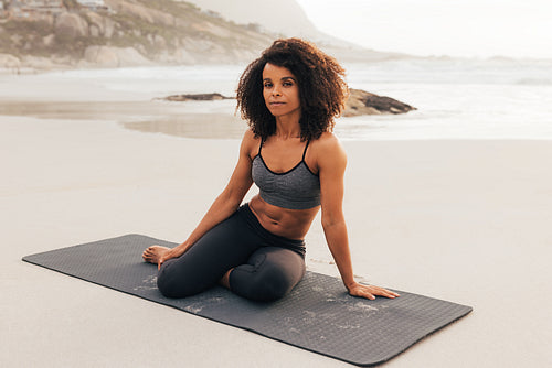 Portrait of a beautiful female sitting on a mat on beach. Woman relaxing after yoga exercises at sunset looking at camera.