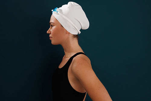 Side view of a woman swimmer standing at blue wall, wearing swimming cap and glasses