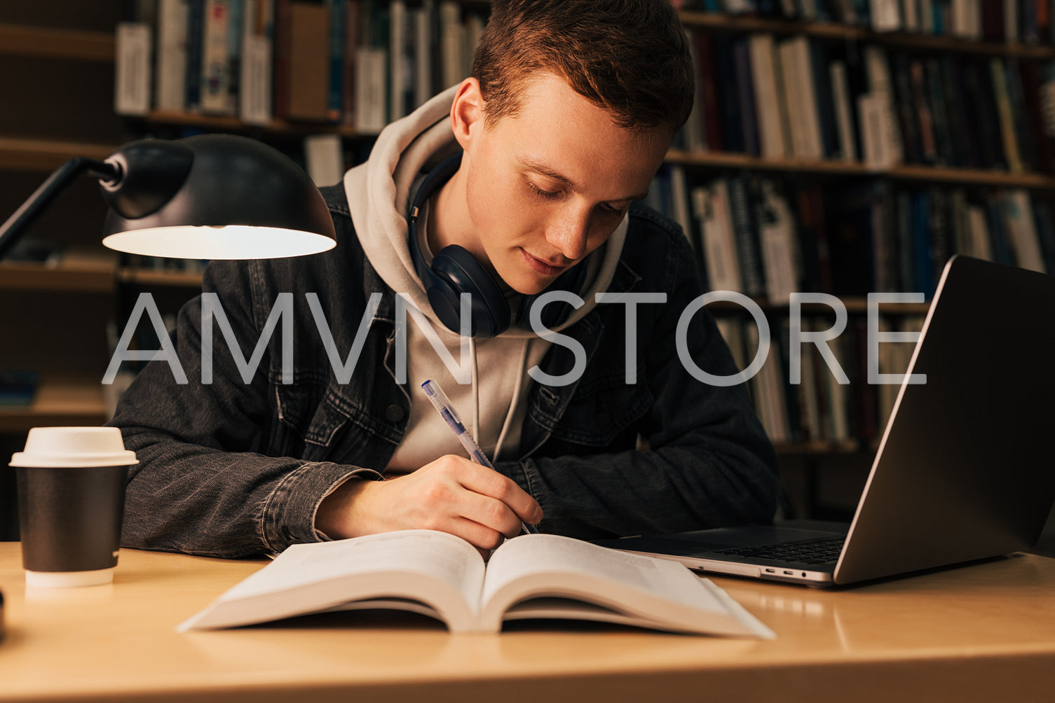 Young man writing while sitting at desk with laptop in library