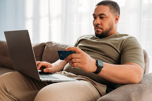 Young man sitting on sofa in living room and making online purchases with credit card