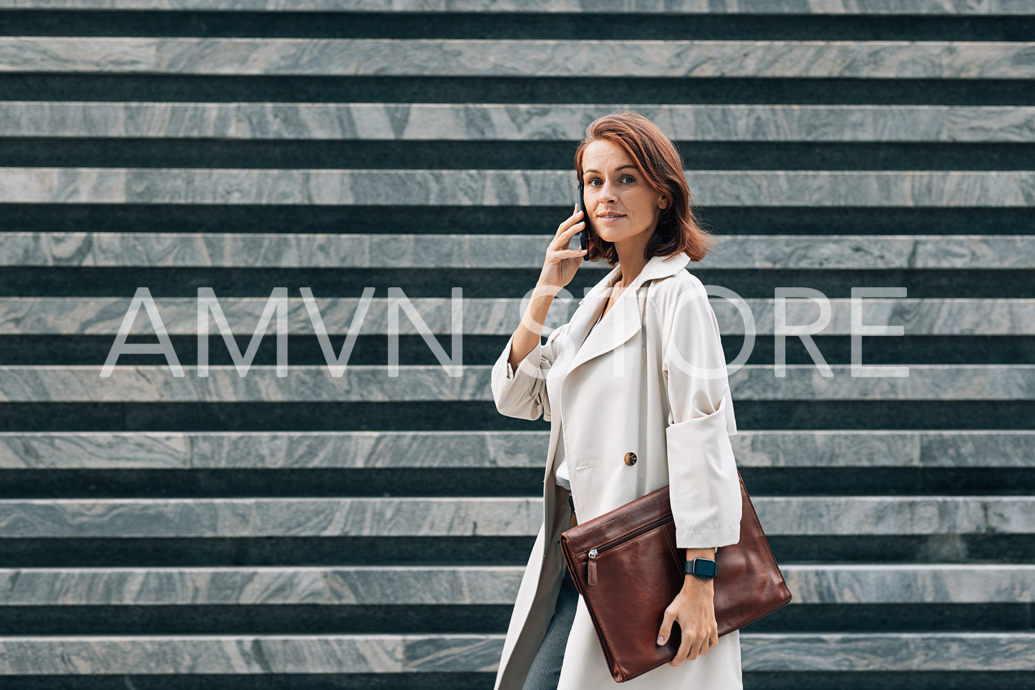 Side view of stylish middle-aged businesswoman in coat. Middle-aged female talking on mobile phone while walking.