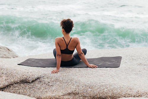 Rear view of young muscular woman sitting on yoga mat and looking on waves